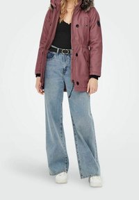 Parka bordeaux avec capuche doublée de fourrure, poignets noirs, fermeture éclair et détails à boutons ; associée à un jean large bleu clair et des baskets blanches.