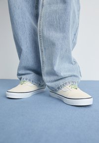 Beige canvas sneakers met groene geblokte accenten, zwarte randen en witte rubberen zolen, gecombineerd met losse lichtblauwe denim broeken.