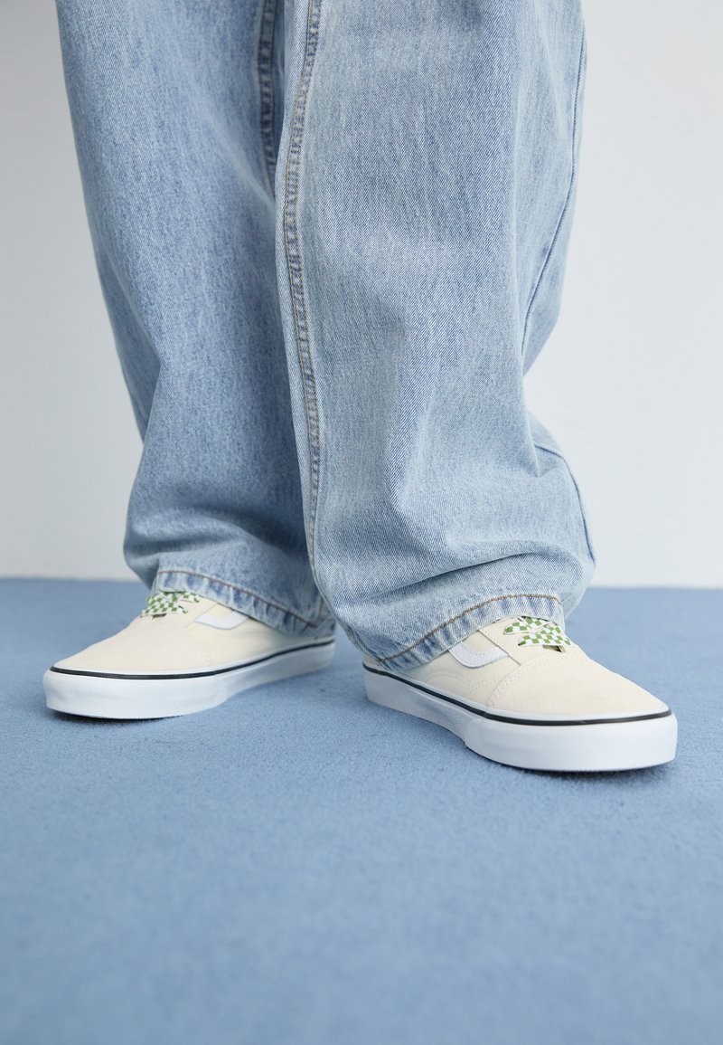 Beige canvas sneakers met groene geblokte accenten, zwarte randen en witte rubberen zolen, gecombineerd met losse lichtblauwe denim broeken.