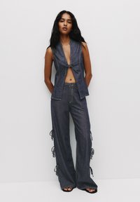 Mörk denim tvådeladชุด som har en ärmlös topp med knytframskärning och vida byxor med sidoknytning. Slät textur och kontrasterande sömnadsdetaljer.
