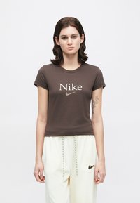 Καφέ βαμβακερό t-shirt με κοντά μανίκια και λογότυπο «Nike» σε ανοιχτό μπεζ χρώμα. Φορεμένο με παντελόνια κρεμ, παρουσιάζοντας μίνιμαλ σχεδιασμό.