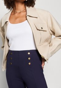 béžová bunda s kratším střihem, velkým límcem a přední kapsou. Pod tím bílý žebrovaný top. Námořnické kalhoty se zlatými knoflíkovými detaily.