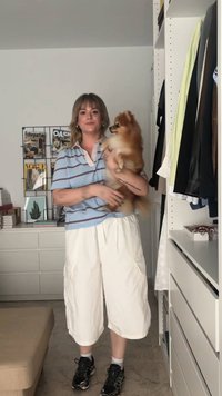 Polo de color azul claro con rayas rojas, pantalones cargo blancos y deportivas negras. Un pequeño Pomerania está sostenido en los brazos del sujeto.