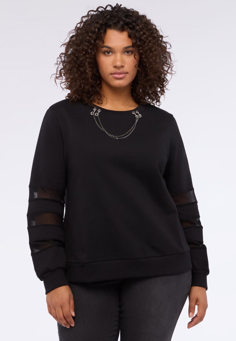 Mujer con cabello rizado que lleva una sudadera negra con paneles de manga transparentes y detalles en cadena cerca del escote, de pie contra un fondo blanco.