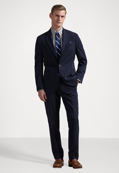 Abito pinstripe blu navy con giacca monopetto, due tasche frontali e mocassini marroni. Accentuato con una cravatta a righe blu e bianche e un fazzoletto da taschino.