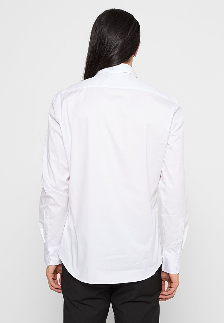 Chemise blanche à manches longues en tissu lisse, avec un col classique, des poignets boutonnés et une coupe ajustée. La vue de dos montre la longueur et la coupe de la chemise.