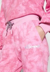 Osoba w różowych dresowych spodniach w tie-dye z dzianym paskiem i białym logo "ellesse", z ręką w kieszeni, dopasowana do podobnej bluzy.