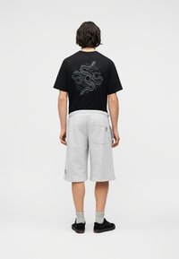 Άτομο με σκούρα μαλλιά που φοράει μαύρο μπλουζάκι με λευκό γραφικό φίδι στην πλάτη, ανοιχτό γκρι σορτς μέχρι το γόνατο, γκρι κάλτσες και μαύρα αθλητικά παπούτσια.