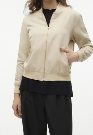 Vrouw draagt een beige jas met ritssluiting, geribbelde manchetten en zoom, zwart topje eronder, en zwarte broek, hand in jaszak.