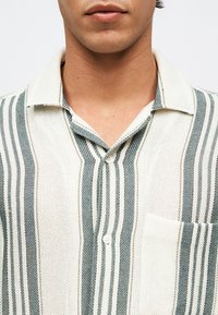 Άνδρας που φοράει πλεκτό πουκάμισο με κατακόρυφες ρίγες σε κρεμ και πράσινο, με γιακά, κουμπιά και τσέπη στην αριστερή πλευρά του στήθους.