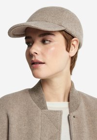 Ljusbrun filtkeps med en rundad skärm, slät textur och sydda detaljer, bärs tillsammans med en beige bomberjacka med ribbad krage.