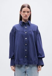 Πουκάμισο από τζιν σε σκούρο μπλε χρώμα, με κούμπωμα με κουμπιά, ευρύ άνετο σχέδιο, μυτερό γιακά και μακριά μανίκια με χαλαρή δομή.