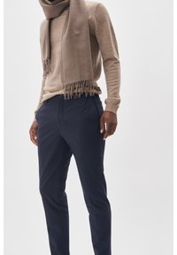 Beige ulltröja med långa ärmar och rundad hals, i kombination med marinblå byxor. En matchande beige scarf med fransar hänger runt halsen.