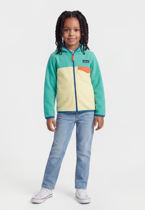 Dieťa s vrkočmi, oblečené v tyrkysovo-krémovej mikine so zipsom a oranžovým vreckom, svetlomodré džínsy a biele tenisky, stojí.