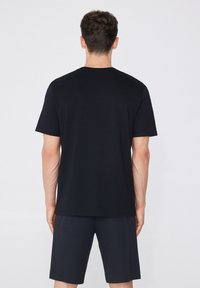Camiseta negra de manga corta con cuello redondo y textura suave, combinada con pantalones cortos oscuros, vista desde atrás.
