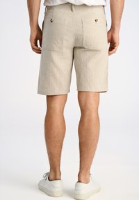 Pantalones cortos rayados beige con una tela texturizada, dos bolsillos traseros con botones y un corte recto, acompañados de zapatillas blancas.