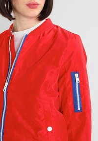 Blouson aviateur rouge en tissu brillant, avec fermeture éclair à l'avant et accent de fermeture éclair bleu sur la poche de la manche. Col côtelé et détail à bouton-pression.