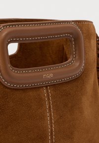 Bolso de ante marrón con un asa de cuero marrón lisa. Presenta costuras tonales y el nombre de la marca 'maje' grabado cerca del asa.