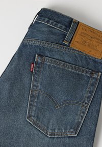Πίσω τσέπη μπλε τζιν Levi's με πορτοκαλί ραφές, κόκκινη ετικέτα Levi's και καφέ δερμάτινο λογότυπο στη μέση.