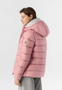 Femme portant une doudoune rose matelassée à capuche avec un intérieur gris clair, se tenant de profil contre un fond blanc uni.