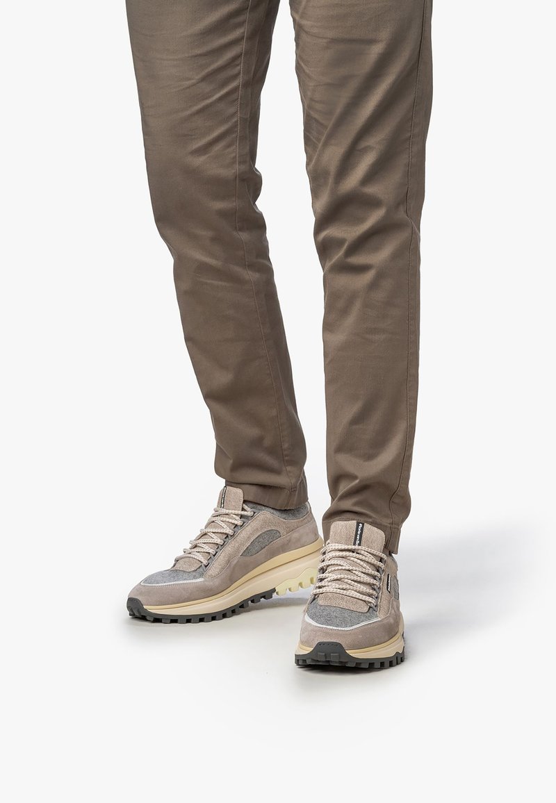 Uomo che indossa pantaloni slim-fit marroni e sneakers beige-grigie con lacci e suole robuste, in piedi su uno sfondo bianco.