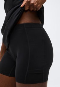 Άτομο σηκώνει μαύρο πουκάμισο για να αποκαλύψει μαύρα σορτς ποδηλασίας ψηλής μέσης με ορατή ραφή σε ανοιχτό φόντο.