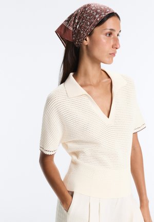 Femme en haut court en maille crème avec col et bordures à motifs, portant un foulard marron à fleurs, posant les mains dans les poches.