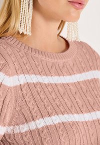Rosa gestrickter Pullover mit weißen horizontalen Streifen, ausgestattet mit einem Zopf-Muster und geripptem Kragen. Ein Paar lange Perlenohrringe sind sichtbar.