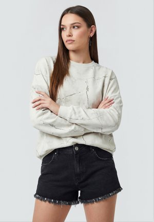 Ung kvinde med langt brunt hår, iført en lys sweatshirt med marmormønster og sorte denimshorts med frynser, armene over kors, kigger til siden.