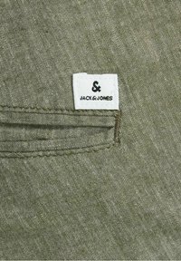 Kieszeni z tkaniny khaki z białą, tkana metką z napisem „JACK & JONES” i symbolem ampersanda, ozdobiona przeszytym obramowaniem.