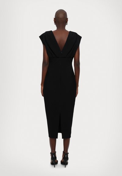 Black fitted dress with a deep V-back, cap sleeves, and a back slit, made of smooth fabric. Heels accentuate the look.