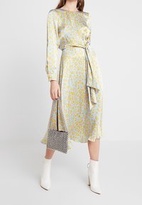 Vaaleansininen midi-mekko, jossa on keltaisia leopardipilkkuja, pitkät hihat, sidottava vyö ja virtaava siluetti. Yhdistetty valkoisiin nilkkureihin ja musta-valkoiseen kuviolliseen laukkuun.