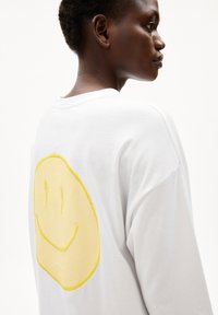 T-shirt bianco in cotone con vestibilità rilassata, caratterizzato da una grande stampa di un volto sorridente giallo sul retro. Maniche corte e scollo tondo.