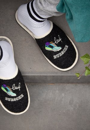 Black felt slippers with white trim, featuring colorful embroidered sneaker design. Ankle-height white socks with black stripes are worn.