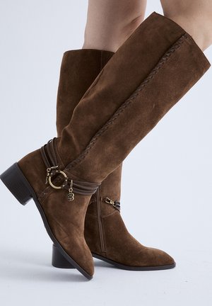 Botas marrones de ante hasta la rodilla con tacones bajos tipo bloque, correas decorativas envolventes y cierres de cremallera, usadas en las piernas contra un fondo liso.