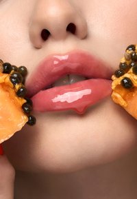 Zbliżenie na błyszczące usta, lśniące wilgocią, otoczone jaskrawym pomarańczowym owocem i ciemnymi ziarnami, ukazujące gładką teksturę.