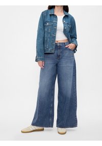 Chaqueta de mezclilla en azul claro, con cierre de botones y bolsillos en el pecho. Combinada con jeans de pierna ancha en azul y zapatillas deportivas blancas.