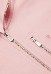 Sudadera rosa con cremallera y cremalleras plateadas, con una textura suave y un detalle de logo bordado encima de la cremallera izquierda.