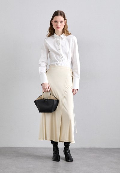 Camicia bianca a maniche lunghe con bottoni, gonna avvolgente crema e borsa nera strutturata. Il modello indossa eleganti stivaletti neri, realizzati con materiali lisci.