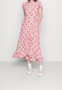 Wickelkleid aus hellrosa Stoff mit floralem Muster, Rüschenärmeln und gestuftem Saum. Kombiniert mit weißen Kniestrümpfen und Turnschuhen.