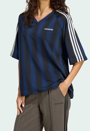 Person iført en navy- og sortstribet Adidas-trøje med hvide ærmestriber og grå Adidas-bukser med sorte sidestriber, hånd i lommen.