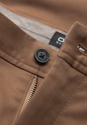 Pantalones de algodón marrón con un botón negro, cierre de cremallera y una sutil textura diagonal. Etiqueta visible cerca de la cinturilla.