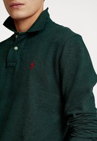 Dunkelgrünes Poloshirt aus strukturiertem Stoff, mit umschlagbarem Kragen, zwei Knöpfen und einem kleinen roten Logo auf der linken Brust.