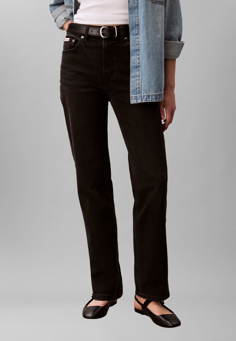 Άτομο που φοράει μαύρο τζιν με ευθείες γραμμές και μαύρη ζώνη, λευκή μπλούζα, τζιν μπουφάν με ρυθμισμένα μανίκια και μαύρα επίπεδα παπούτσια με λουράκια.