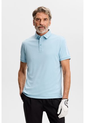 Ældre mand med gråt hår og skæg iført en lyseblå polo og sorte bukser, med en hvid golfhandske på venstre hånd.