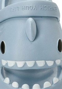 Chancla de goma de color azul claro diseñada como un tiburón, con una boca sonriente y dientes blancos, y grandes ojos negros. El texto dice "ENRIQUECE TU VIDA."