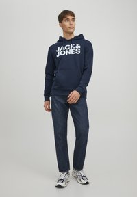 Navy hoodie with white "JACK & JONES" logo, front pocket, pullover design, paired with dark blue jeans and white/black sneakers.