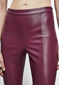 Pantaloni di pelle bordeaux con una texture liscia, dotati di vita alta e cuciture precise sul davanti, con una chiusura a zip sul lato.