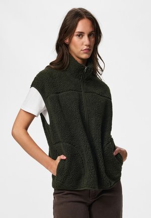 Jeune femme portant un gilet polaire zippé vert foncé avec des poches par-dessus un t-shirt blanc à manches courtes, debout les mains dans les poches.
