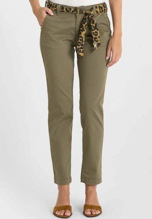 Vrouw draagt olijfgroene smalle broek met een ceintuur met luipaardprint, en bruine sandalen met open tenen, staat met de handen bij de zakken.